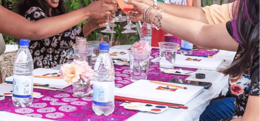 Ladies hands meet in the centre of the table for a celebratory toast, with canvases ready to be painted below them. Faces of the participants are not visible.