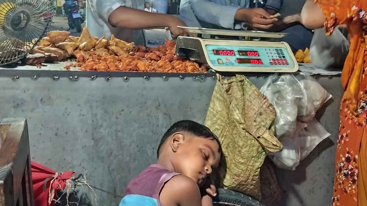 A young boy naps with his head resting on the wheel of a temporary food cart while his mother works, serving food in the evening market. The warm sunset light captures both her struggle and his innocence.