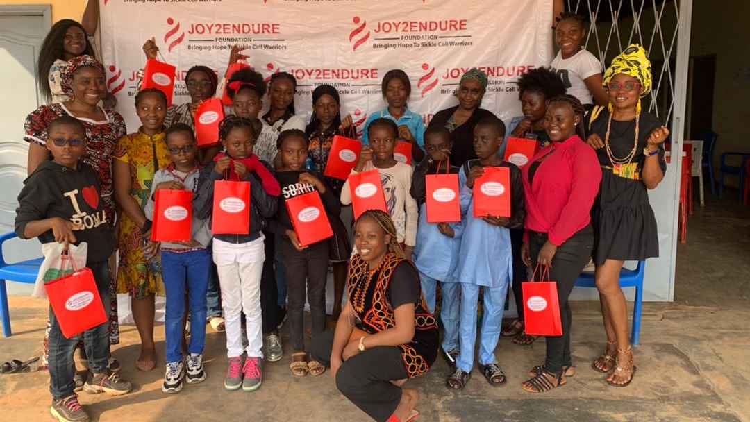 A Family photo taken during our maiden Warrior Empowerment Program (WEP). Warriors stand against a backdrop with the Joy2Endure Foundation Logo, holding branded gift bags containing routine daily medications and hot water bottles.