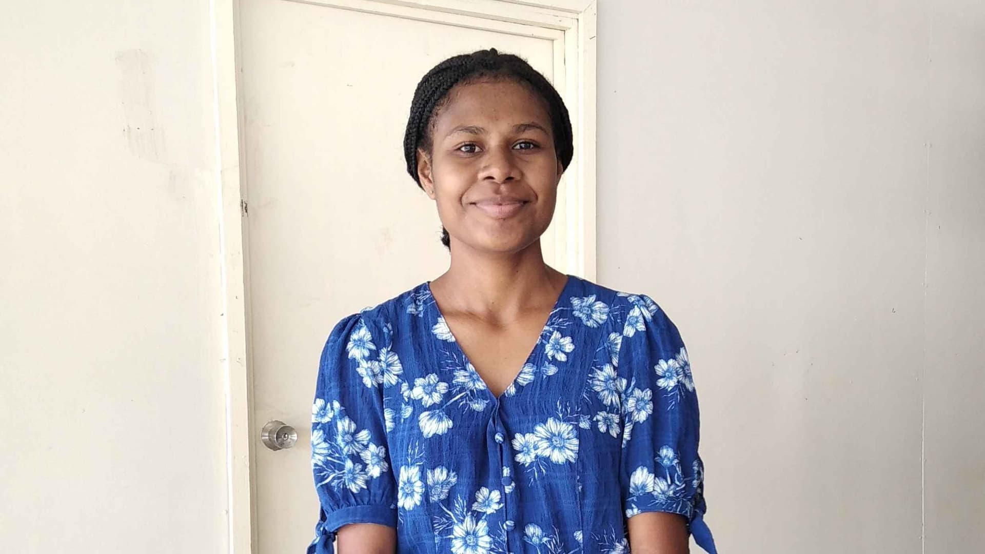 Smiling woman in a blue floral dress stands in front of a light-colored door. The scene is simple and serene, conveying a warm, welcoming vibe.