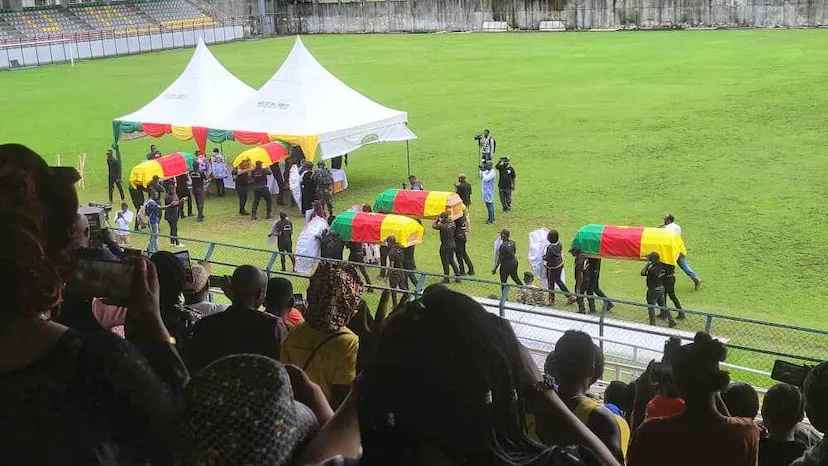 Pallbearers carry five caskets draped in the flag of Cameroon as funeral attendees look on from stadium seats.
