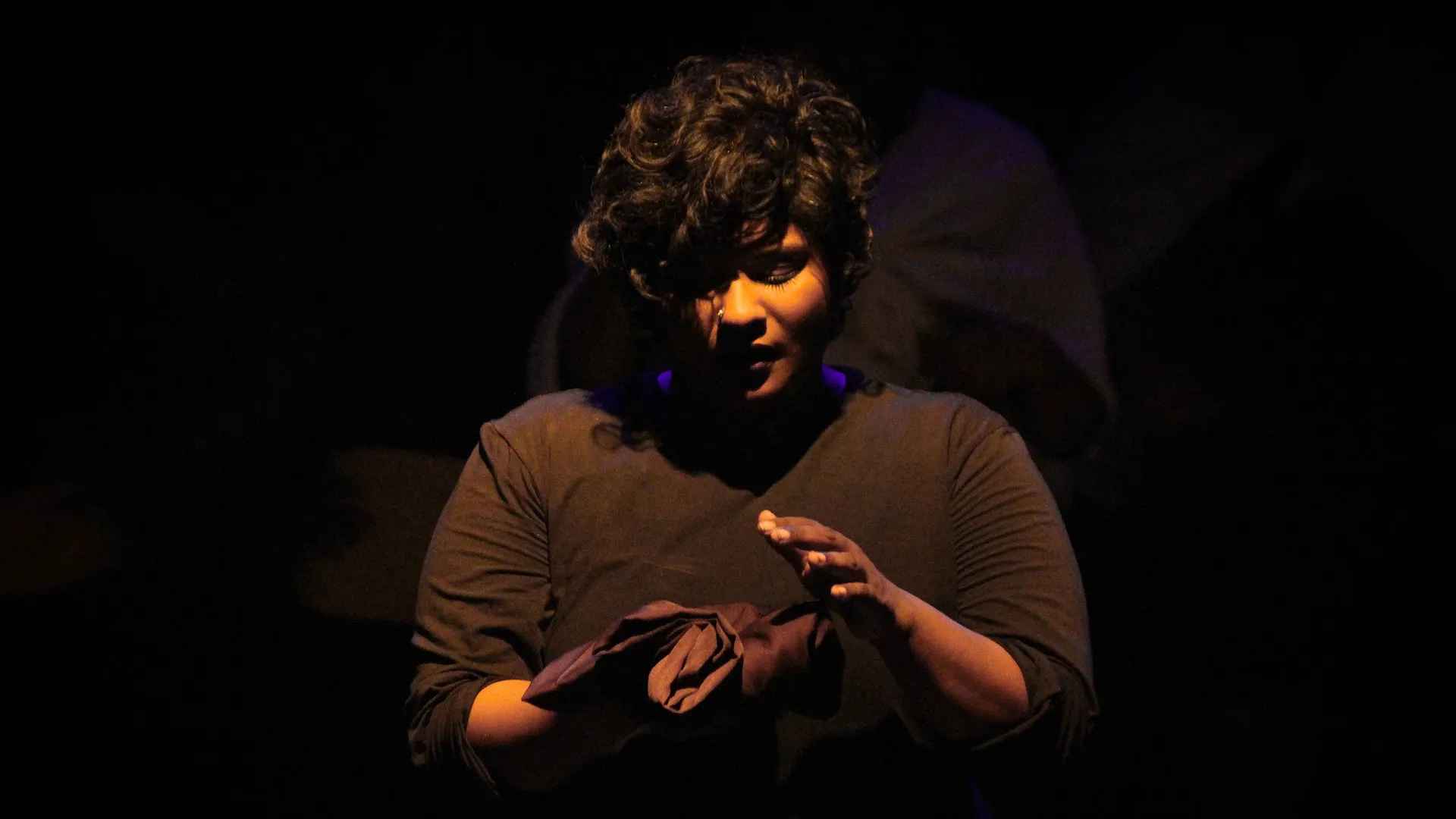 A woman with short dark curly hair looks down at her hands while holding a piece of fabric. She is surrounded by a dark backdrop.